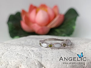 The Angelic Marquise Genuine Birthstones Ring in silver features alternating white and green gemstones set in an elegant design, presented on a textured stone surface with a soft-focus flower background.