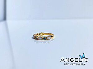 A gold ring featuring three gemstones and two white stones, displayed on a white surface. The brand name 'Angelic DNA Jewellery' is visible at the bottom right corner of the image.