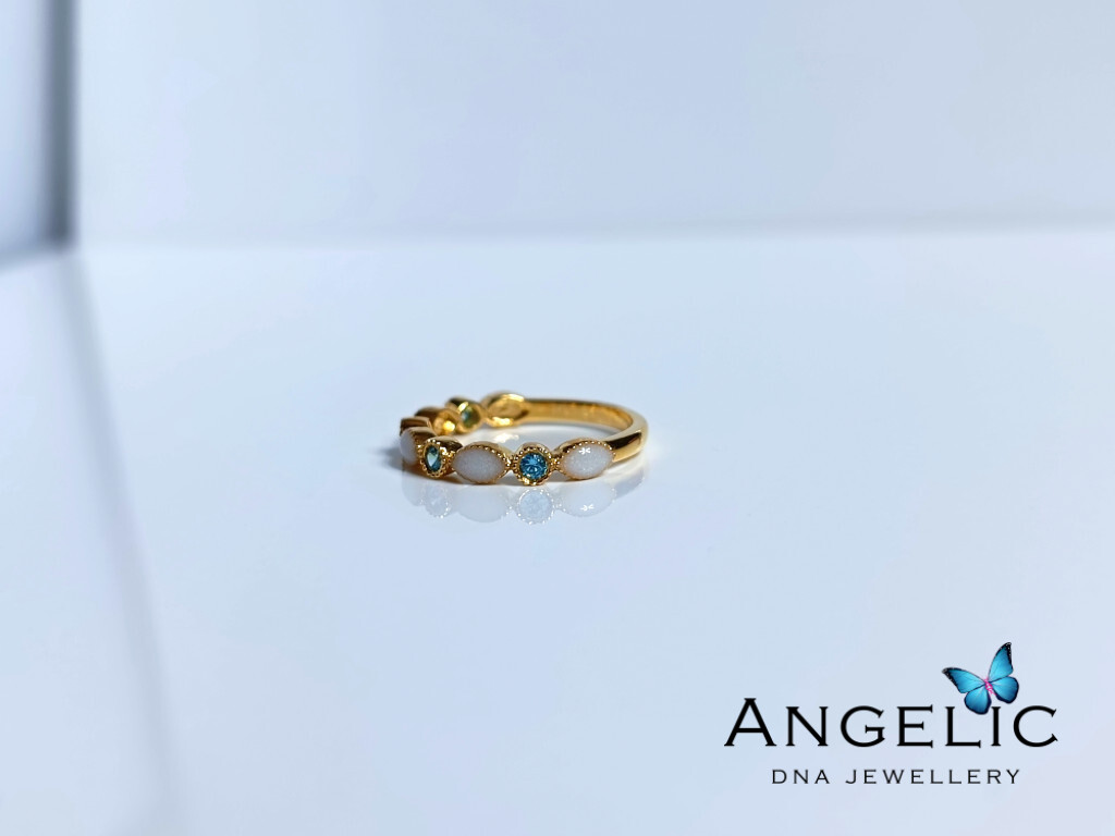 A gold ring featuring three gemstones and two white stones, displayed on a white surface. The brand name 'Angelic DNA Jewellery' is visible at the bottom right corner of the image.