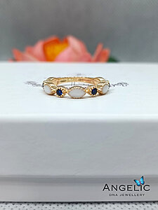 A gold ring featuring white opal and blue stone accents, displayed on a white jewelry box with the 'Angelic DNA Jewellery' logo visible.