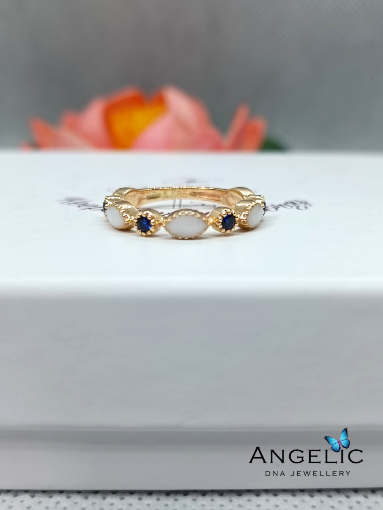 A gold ring featuring white opal and blue stone accents, displayed on a white jewelry box with the 'Angelic DNA Jewellery' logo visible.