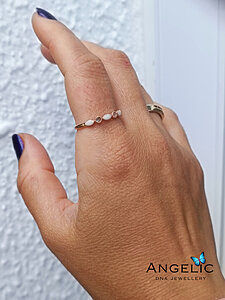A close-up of a woman's hand displaying a delicate gold ring with small stones, set against a textured white wall. Another ring is seen on the same hand, and the nails are painted dark blue.