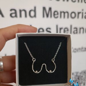 The Angelic Boobies Necklace (Handmade Limited Edition) features a delicate silver chain with a unique design resembling two abstract shapes. It is presented in a simple white box with a black insert.