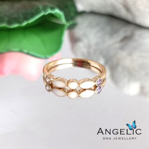 Gold ring with intricate design featuring white enamel and purple accents, displayed on a soft surface with green foliage in the background, promoting Angelic DNA Jewelry.