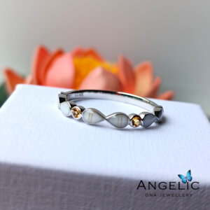 Silver infinity bracelet with gold accents displayed on a white pedestal, surrounded by soft pink flowers, showcasing elegant jewelry design by Angelic.