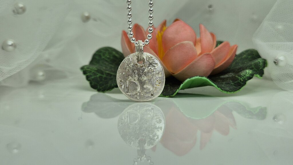 Silver pendant necklace with intricate floral design, displayed in front of a pink lotus flower, reflecting light on a smooth surface.