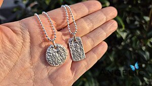 Hand holding two unique silver pendants, one round and one rectangular, both featuring textured designs, suspended from ball chain necklaces.